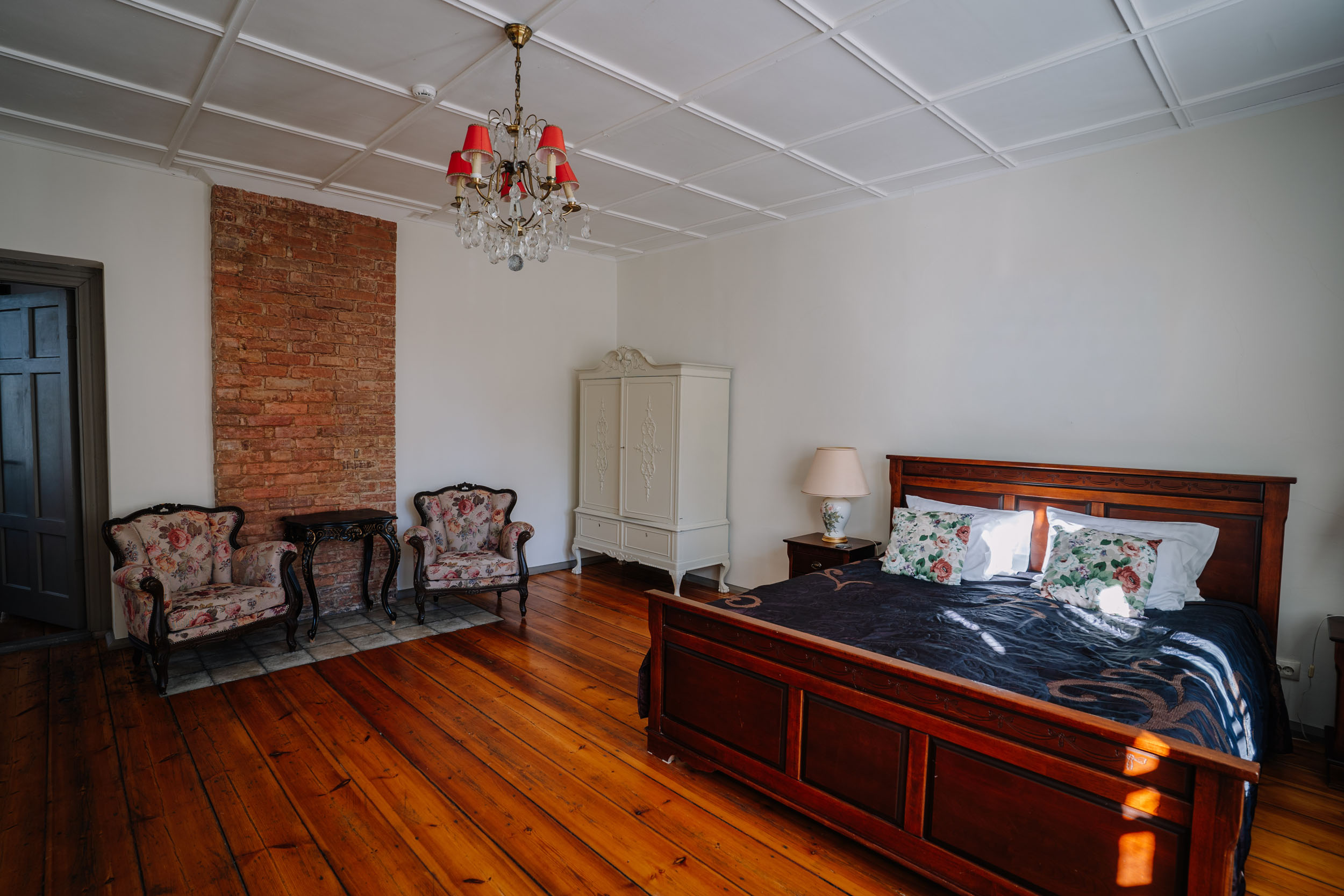 Cozy bedroom showcasing a wooden bed with floral bedding, vintage armchairs, a chandelier, and a brick accent wall, illuminated by natural light